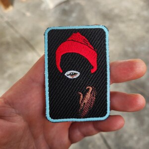 Pode incluir: Patch bordado com fundo preto, formato curvo vermelho e design de olho. O patch tem uma borda azul clara. O design também inclui uma forma marrom. O patch é retangular.