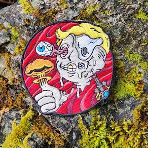 May include: Embroidered patch featuring a cartoon character with a detached eyeball and a mushroom cloud. The design includes a yellow hairstyle, a red background, and a black border. The character is holding a miniature mushroom cloud.
