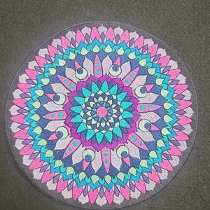 May include: Circular mandala design in shades of pink, purple, teal, and blue on a dark gray background. The intricate pattern features symmetrical floral and geometric elements, creating a visually appealing decorative item.