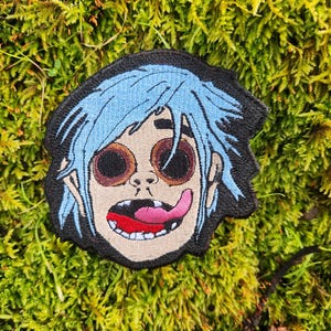May include: Embroidered patch featuring a cartoon character with light blue hair, large brown eyes, and a pink tongue sticking out. The patch has a black border and is set against a background of green moss.