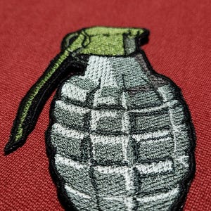 Op de afbeelding: Geborduurde patch van een handgranaat. De patch heeft een grijze behuizing met een rasterpatroon, een groene bovenkant en een zwarte handgreep. De patch staat op een rode achtergrond.