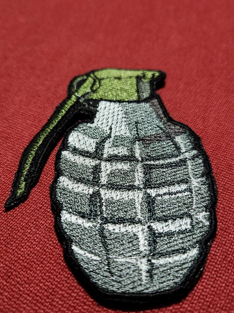 Op de afbeelding: Geborduurde patch van een handgranaat. De patch heeft een grijze behuizing met een rasterpatroon, een groene bovenkant en een zwarte handgreep. De patch staat op een rode achtergrond.