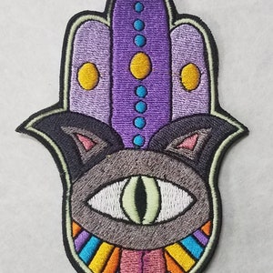 Puede incluir: Parche bordado con forma de mano de Hamsa con diseño de cara de gato. El parche presenta una cara de gato gris con un ojo, un diseño morado y amarillo y una lengua colorida. El parche está delineado en negro.