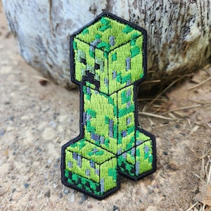 May include: An embroidered patch featuring a pixelated green creature with a blocky head and body. The design uses shades of lime green, dark green, and gray thread to create a textured, square-patterned appearance, all outlined in a thick black border.
