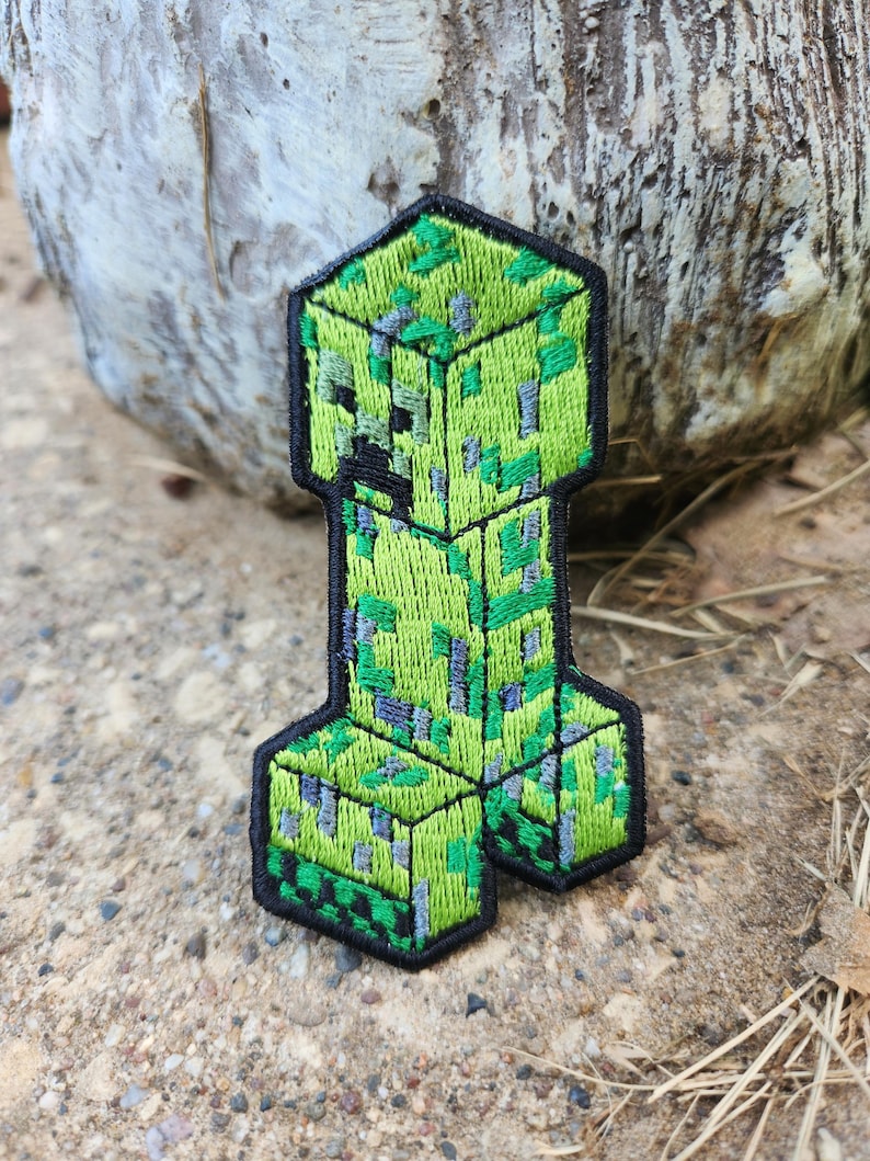 K&ouml;nnte beinhalten: Ein gestickter Aufn&auml;her, der eine verpixelte gr&uuml;ne Kreatur mit blockartigem Kopf und K&ouml;rper zeigt. Das Design verwendet verschiedene Nuancen von Limettengr&uuml;n, Dunkelgr&uuml;n und Grau, um eine texturierte, quadratische Optik zu erzeugen, eingefasst von einem dicken schwarzen Rand.