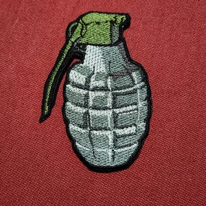 Op de afbeelding: Geborduurde patch van een grijze handgranaat met een groene bovenkant en zwarte trekpen. De patch staat op een rode achtergrond. De granaat heeft een gedetailleerd, gestructureerd ontwerp.
