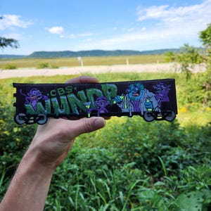 May include: A rectangular embroidered patch featuring a black background with the green text WUNDR and CBS. The design includes purple monkey-like figures and a blue-skinned character holding yellow test tubes, all set against a train car outline.