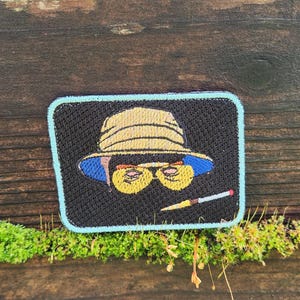May include: Embroidered patch depicting a cartoon character in a hat and sunglasses, with a cigarette. The patch has a black background with a light blue border. The hat is yellow, and the sunglasses are yellow with a blue rim.