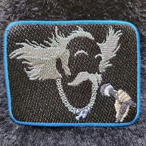 May include: Embroidered patch featuring a stylized, cartoonish face with a long mustache and a chain around the neck. The patch has a black background with a blue border and is set against a dark, textured surface.