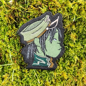 May include: Embroidered patch featuring a green-skinned character with black hair and a hat. The patch has a black border and is set against a background of green moss. The character's design includes details in white, green, and gold.