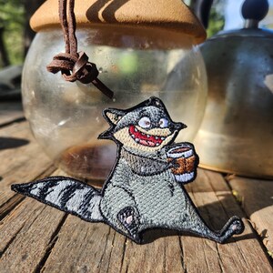 Op de afbeelding: Geborduurde patch met een cartoon wasbeer die een koffiemok vasthoudt. De wasbeer is grijs met zwart-witte strepen op zijn staart. De patch heeft een rode mond. Vage achtergrond met koffieapparatuur.