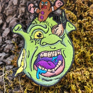 May include: Embroidered patch featuring a green ogre with a wide open mouth, purple tongue, and blue drool. A gingerbread man is perched on the ogre's head. The patch has black edging and is set against a textured background.