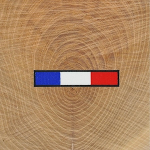 Peut inclure: Un écusson rectangulaire avec le drapeau français. L'écusson présente des rayures horizontales bleues, blanches et rouges, soulignées de noir. L'écusson est posé sur un fond brun clair, veiné de bois.
