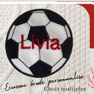 Puede incluir: Parche bordado de balón de fútbol en blanco y negro con el nombre "Livia" en rojo. El parche está sobre un fondo de punto color crema. El texto en la parte inferior dice "Ecusson brodé personnalisé Choix multiples."