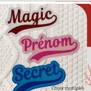 Peut inclure: Trois patchs brodés thermocollants avec un dos en tissu blanc. Les patchs ont la forme d'une bannière incurvée. Le patch du haut est rouge avec le mot "Magic" en blanc. Le patch du milieu est rose avec le mot "Prénom" en blanc. Le patch du bas est bleu avec le mot "Secret" en blanc. Les patchs sont sur un fond en tissu blanc tricoté.