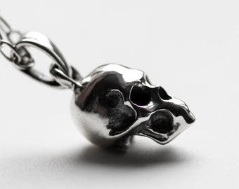 May include: A silver skull pendant necklace with a detailed design. The skull is facing to the side and has a chain attached to the back.