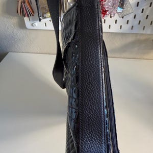 Puede incluir: Bolso de hombro negro con un patr&oacute;n texturizado similar al de un cocodrilo. El bolso tiene una correa larga y ajustable y un asa de cadena plateada. Un bolsillo con cremallera es visible en el lateral. El bolso est&aacute; sujeto por una correa negra.