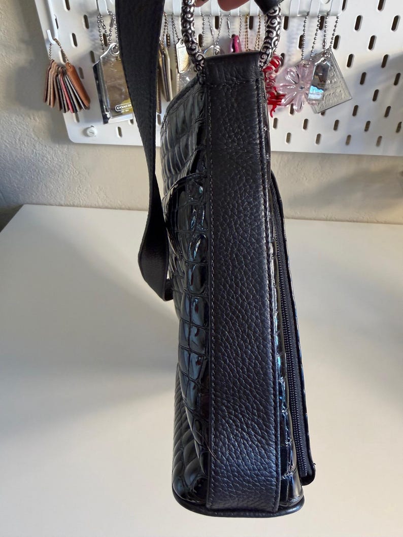 Puede incluir: Bolso de hombro negro con un patr&oacute;n texturizado similar al de un cocodrilo. El bolso tiene una correa larga y ajustable y un asa de cadena plateada. Un bolsillo con cremallera es visible en el lateral. El bolso est&aacute; sujeto por una correa negra.