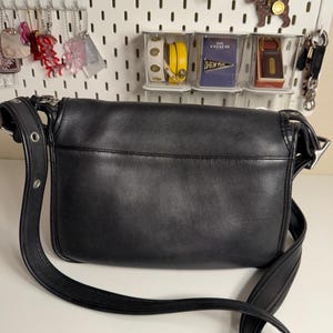May include: Black leather crossbody bag with a long adjustable strap. The bag has a flap closure and silver-tone hardware. The bag is sitting on a white surface. The background includes various accessories hanging on a white pegboard.