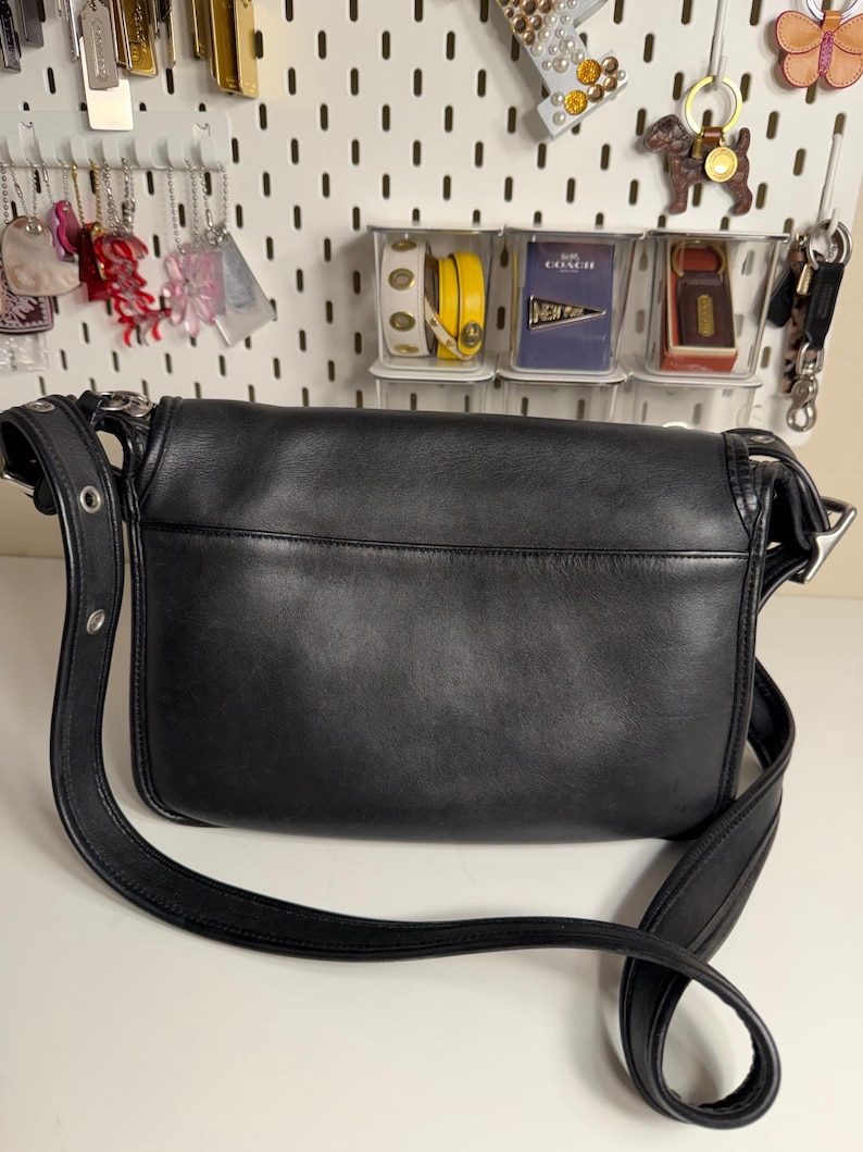 May include: Black leather crossbody bag with a long adjustable strap. The bag has a flap closure and silver-tone hardware. The bag is sitting on a white surface. The background includes various accessories hanging on a white pegboard.