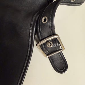May include: Close-up of a black leather bag with a silver-colored buckle and strap. The bag has a stitched edge and a smooth, shiny surface. The strap is attached with metal rivets.
