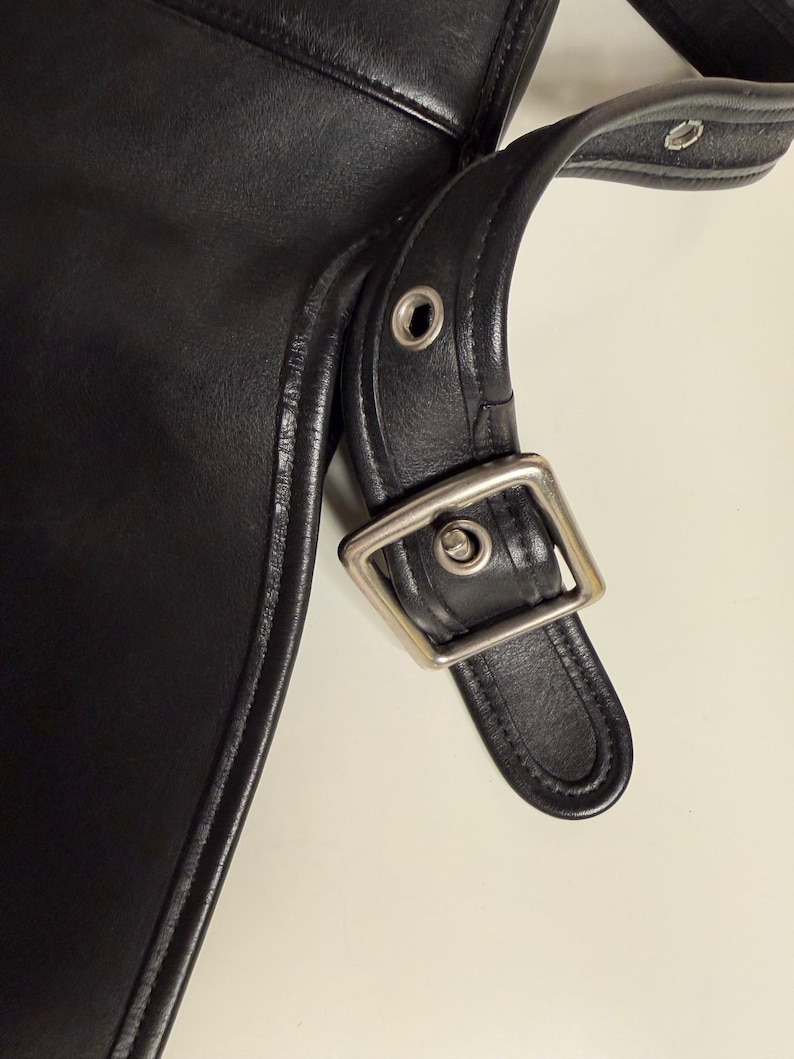 May include: Close-up of a black leather bag with a silver-colored buckle and strap. The bag has a stitched edge and a smooth, shiny surface. The strap is attached with metal rivets.