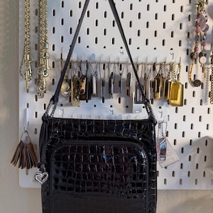 Puede incluir: Bolso de hombro negro brillante con textura de cocodrilo. El bolso tiene un bolsillo frontal y un charm de coraz&oacute;n plateado. La correa negra cuelga de un panel perforado blanco con joyas.
