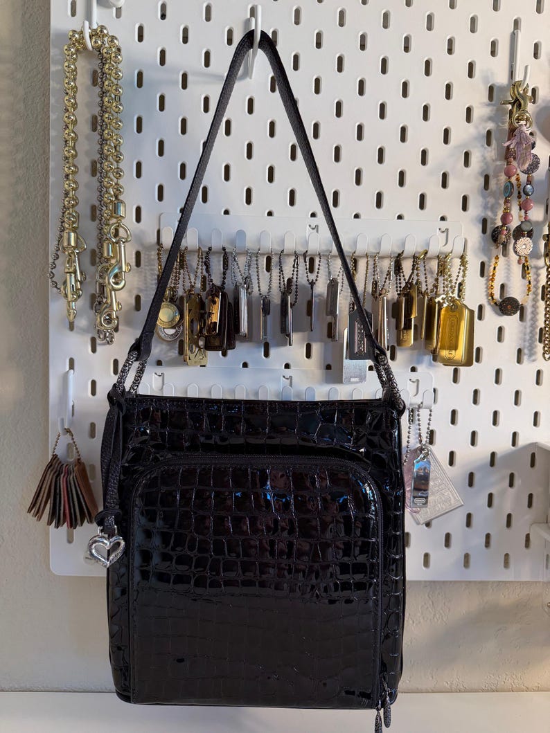Puede incluir: Bolso de hombro negro brillante con textura de cocodrilo. El bolso tiene un bolsillo frontal y un charm de coraz&oacute;n plateado. La correa negra cuelga de un panel perforado blanco con joyas.