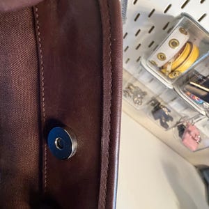 May include: Close-up of a dark brown leather bag with a magnetic closure. The bag's edge is stitched with a lighter brown thread. The interior is a dark brown canvas. The bag is partially visible, with a glimpse of accessories in the background.