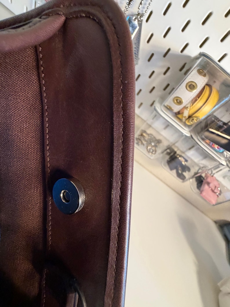May include: Close-up of a dark brown leather bag with a magnetic closure. The bag's edge is stitched with a lighter brown thread. The interior is a dark brown canvas. The bag is partially visible, with a glimpse of accessories in the background.