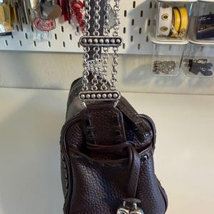 May include: A dark brown leather handbag with a textured finish. The bag features a silver chain strap and a heart-shaped charm. The bag has a small pocket on the front and a drawstring closure.