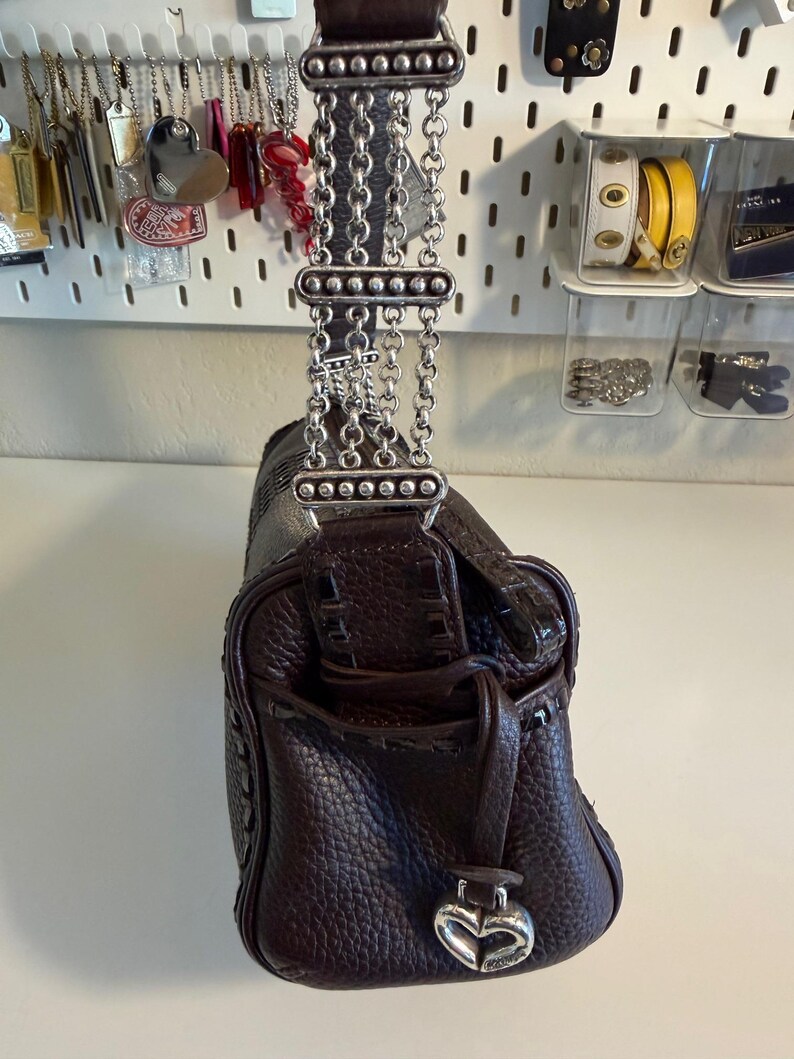 May include: A dark brown leather handbag with a textured finish. The bag features a silver chain strap and a heart-shaped charm. The bag has a small pocket on the front and a drawstring closure.