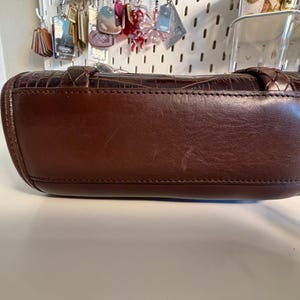 May include: A brown leather handbag with a textured crocodile pattern. The bag has a rounded shape with a stitched seam along the bottom. The handle is braided. The bag is sitting on a white surface.