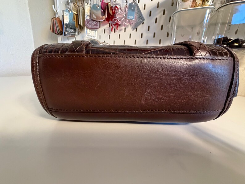 May include: A brown leather handbag with a textured crocodile pattern. The bag has a rounded shape with a stitched seam along the bottom. The handle is braided. The bag is sitting on a white surface.