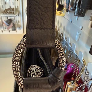 Puede incluir: Bolso de mano de cuero negro con un anillo decorativo de metal plateado y un emblema en forma de coraz&oacute;n. El bolso presenta una correa de cuero texturizado y un detalle de lazo. El bolso est&aacute; colgado de un panel perforado blanco.