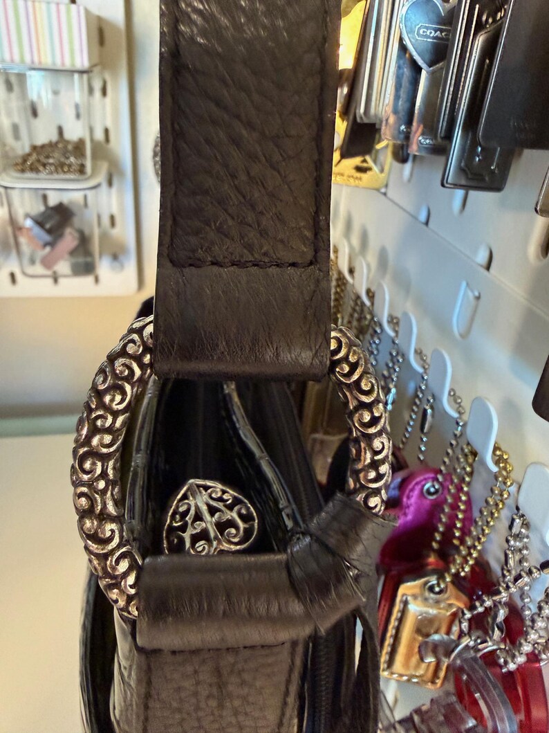 Puede incluir: Bolso de mano de cuero negro con un anillo decorativo de metal plateado y un emblema en forma de coraz&oacute;n. El bolso presenta una correa de cuero texturizado y un detalle de lazo. El bolso est&aacute; colgado de un panel perforado blanco.