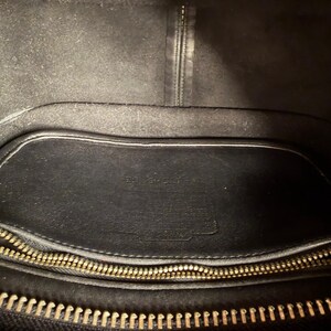 May include: Black leather handbag interior with a gold zipper and a small leather pocket. The interior is lined with a soft, dark material. The bag appears to be well-worn.
