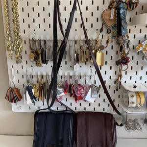 May include: Two rectangular crossbody bags with long adjustable straps, one in black and one in dark brown. Each bag features a flap closure with vertical stitched detailing and a small rectangular metal logo plate centered on the lower front section.