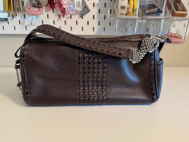May include: A dark brown leather handbag with a woven center panel and a decorative silver chain accent. The bag has a top handle and a long strap for shoulder or crossbody wear. The leather has a textured appearance.