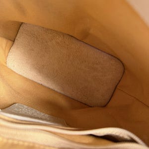 May include: Interior view of a light tan leather bag. The bag's interior is a lighter shade of tan, with a suede-like patch and a small pocket. The stitching is visible along the edges, and the overall design is simple and functional.