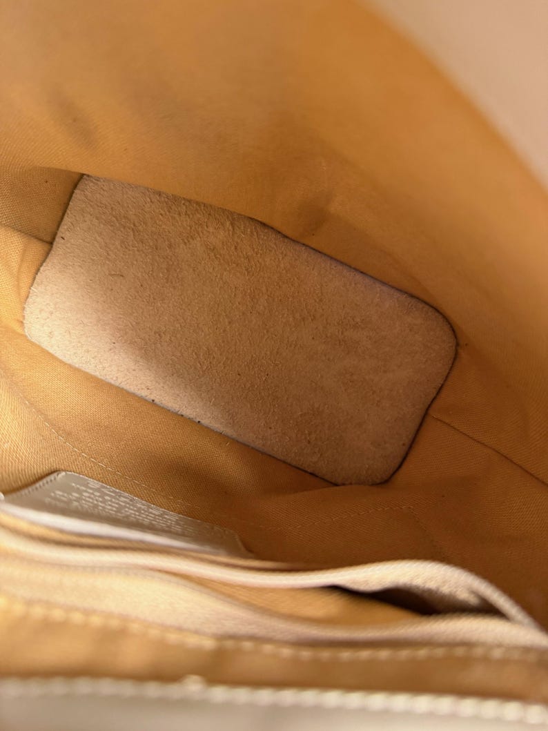 May include: Interior view of a light tan leather bag. The bag's interior is a lighter shade of tan, with a suede-like patch and a small pocket. The stitching is visible along the edges, and the overall design is simple and functional.