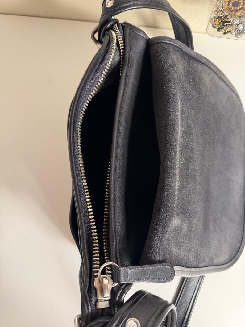 May include: Black leather shoulder bag with silver-tone hardware. The bag features a top handle, a shoulder strap, and multiple compartments. The main compartment is open, revealing the interior. The bag has a zipper closure and a flap closure.