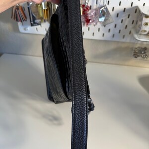 Puede incluir: Bolso de hombro de cuero negro con un patr&oacute;n texturizado similar al de un cocodrilo. El bolso tiene una correa larga y estrecha y un detalle de cadena plateada. El bolso est&aacute; sostenido por una mano.