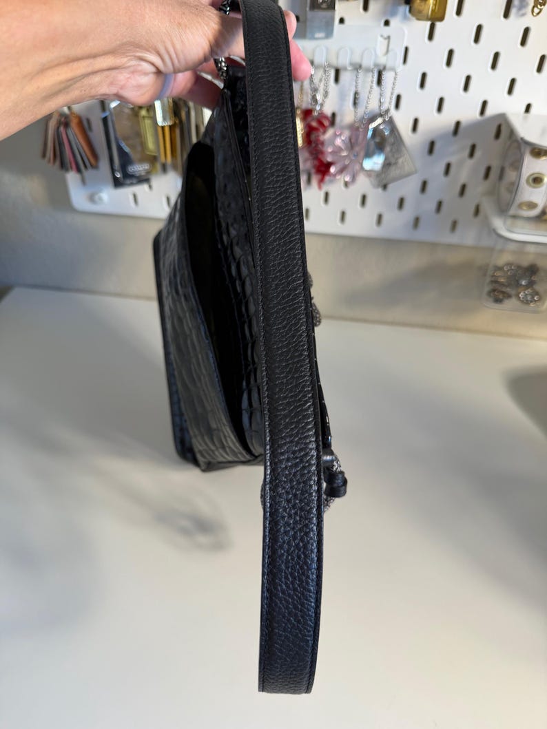 Puede incluir: Bolso de hombro de cuero negro con un patr&oacute;n texturizado similar al de un cocodrilo. El bolso tiene una correa larga y estrecha y un detalle de cadena plateada. El bolso est&aacute; sostenido por una mano.