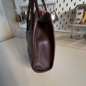 May include: A brown leather tote bag with a braided handle. The bag has a structured shape with a rounded base and a contrasting dark brown trim. The interior is black.