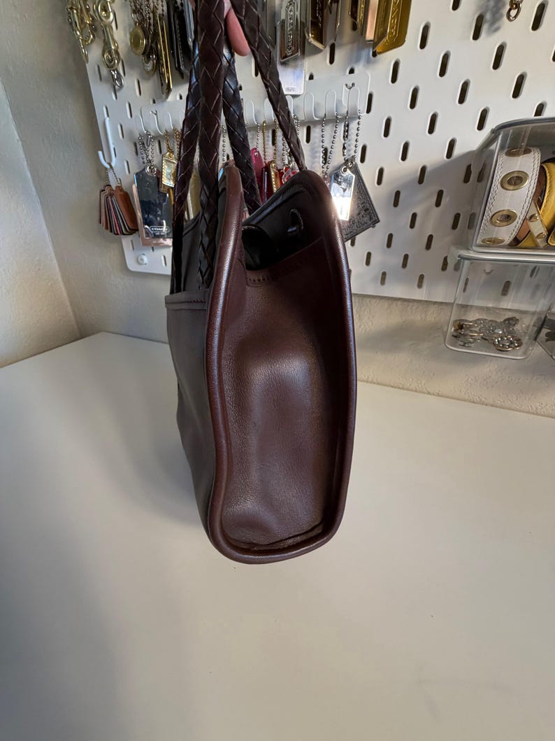 May include: A brown leather tote bag with a braided handle. The bag has a structured shape with a rounded base and a contrasting dark brown trim. The interior is black.