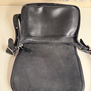 May include: Black leather shoulder bag with a flap closure. The bag has a silver-tone buckle and zipper. The interior is a dark gray suede. The bag has a long adjustable strap.