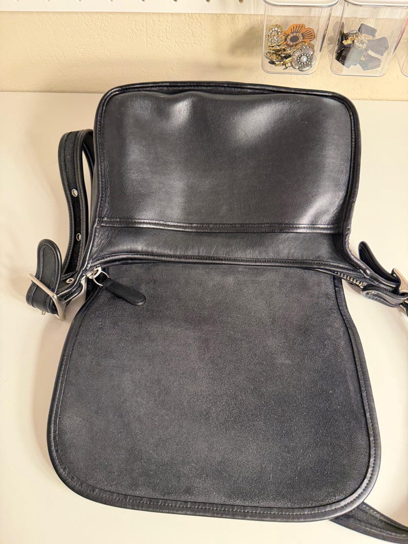 May include: Black leather shoulder bag with a flap closure. The bag has a silver-tone buckle and zipper. The interior is a dark gray suede. The bag has a long adjustable strap.