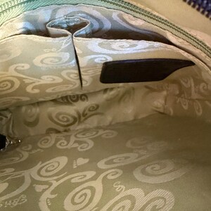 Puede incluir: Vista interior de un bolso negro. El interior presenta una tela verde claro con un dise&ntilde;o de remolino. Se ven varios bolsillos y un cierre de cremallera. Hay una peque&ntilde;a etiqueta de cuero negro.