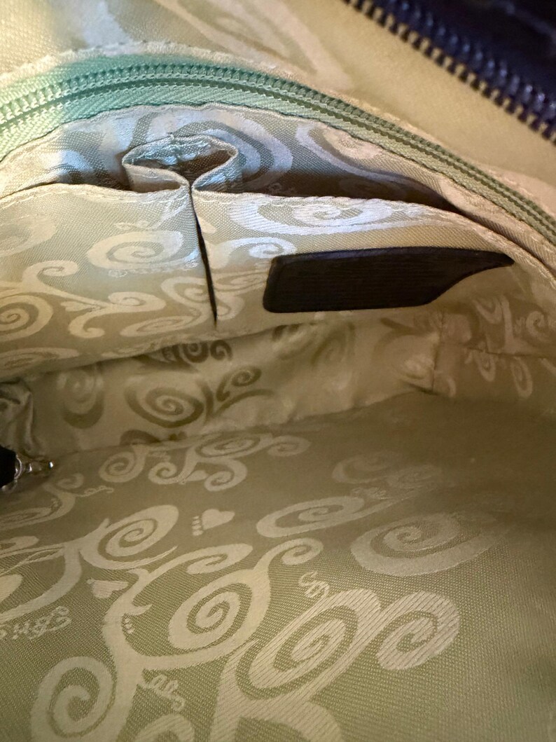 Puede incluir: Vista interior de un bolso negro. El interior presenta una tela verde claro con un dise&ntilde;o de remolino. Se ven varios bolsillos y un cierre de cremallera. Hay una peque&ntilde;a etiqueta de cuero negro.